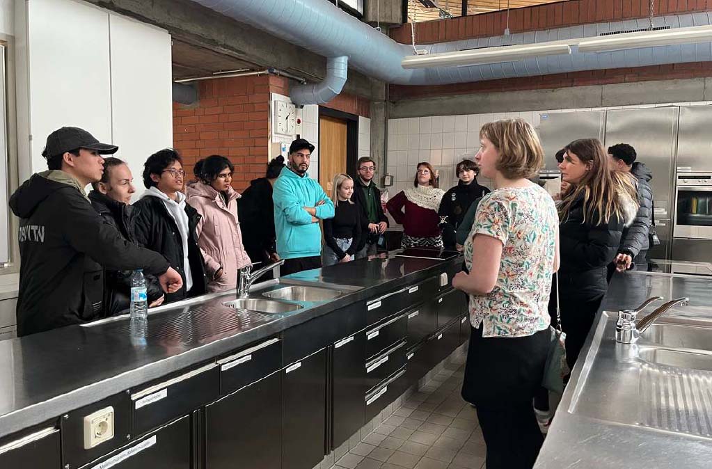 Auszubildende treffen sich zum ersten Mal. Sie stehen in einer Schulküche als Gruppe, eine Lehrerin hält eine Ansprache.